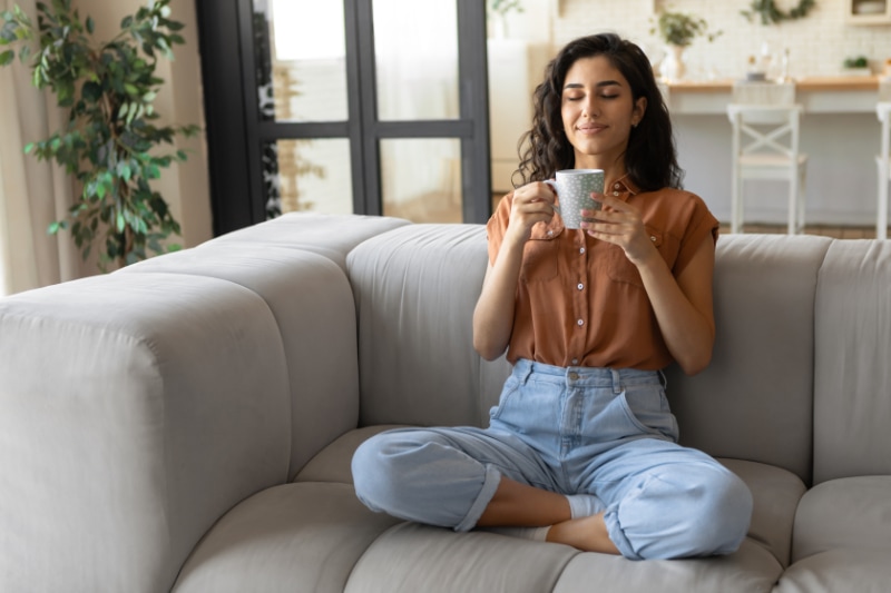 Understanding Your Air Conditioner. Peaceful young brunette woman sitting on couch with closed eyes, drinking hot coffee at home, copy space. Full length of millennial female enjoying lazy morning with warm drink in living room.