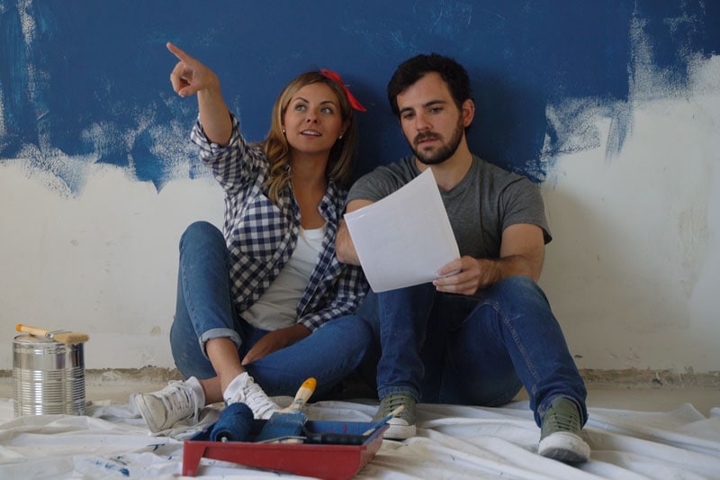 Couple sitting against half painted wall, trying to determine what color to paint it while remodeling their home.