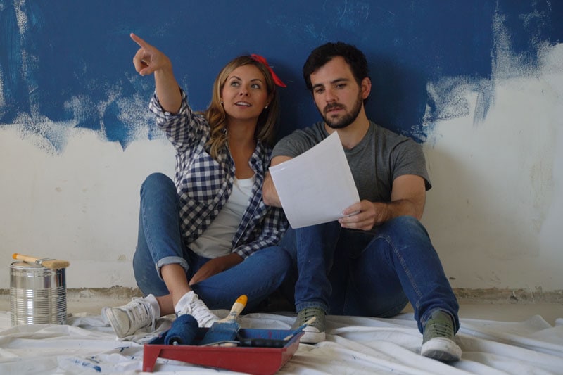 Video - Remodeling Your Home? Don’t Forget Your Comfort! Couple sitting against half painted wall, trying to determine what color to paint it.