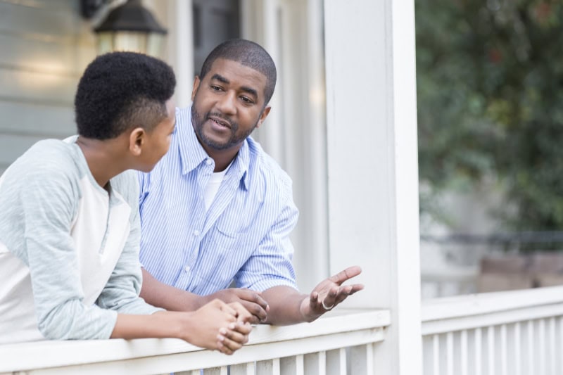 A father leans against the railing of his front porch with his teen son having a serious discussion.