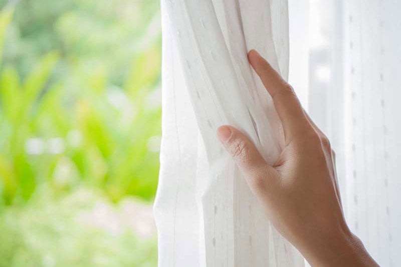 Homeowner opening curtain during spring to represent info about spring money-saving tips.