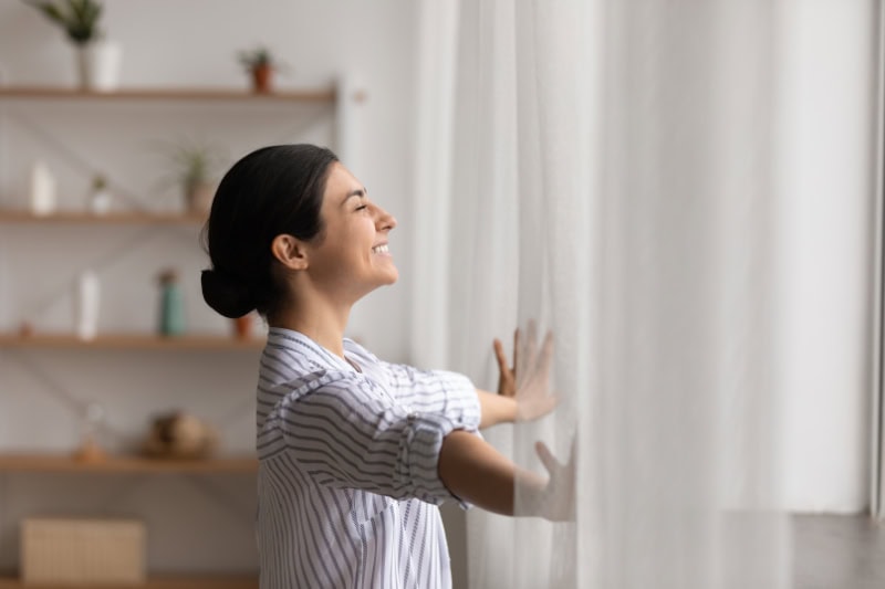 Woman smiling as she opens her curtains to a beautiful day. Representing why ductless is the way to go.